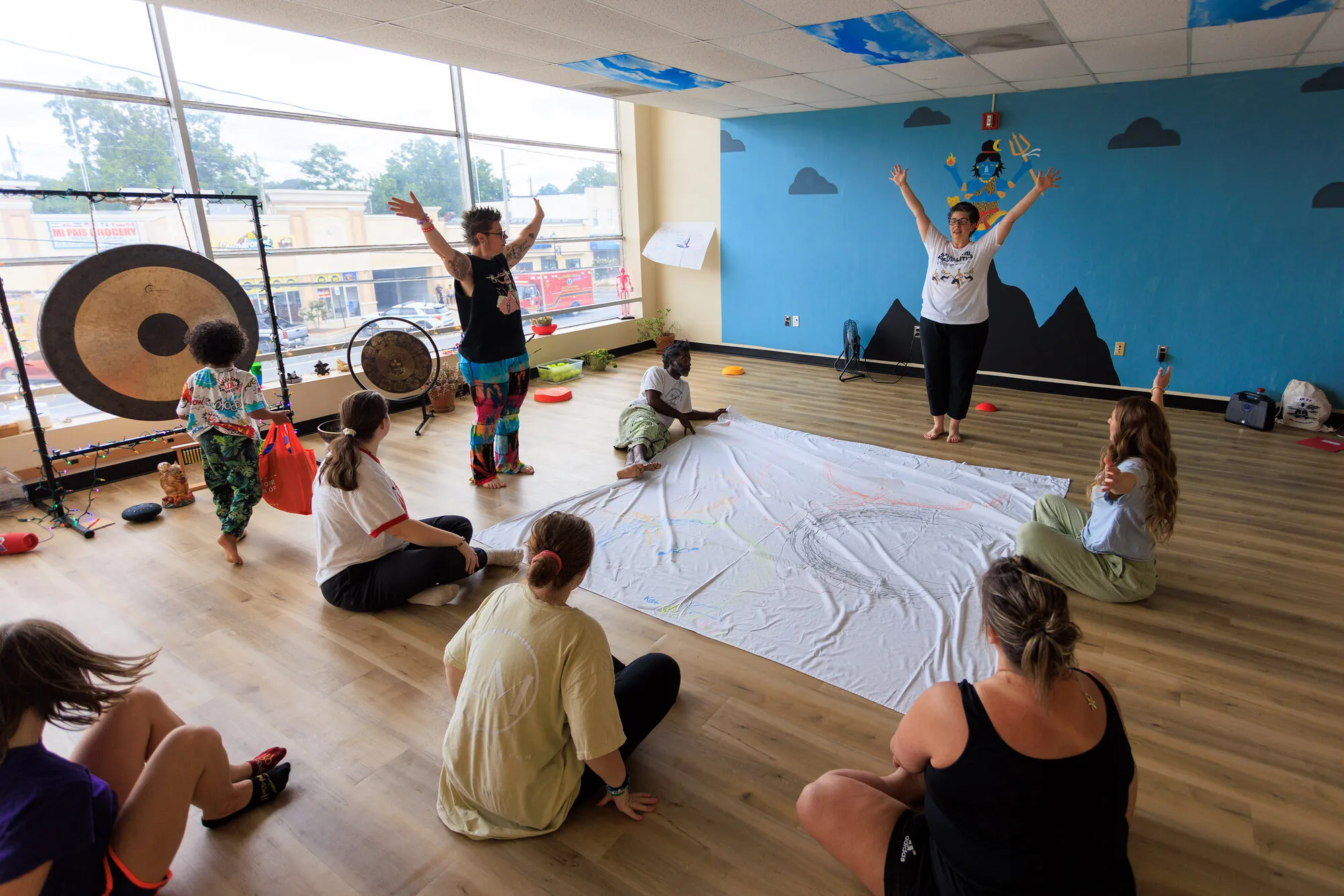 Adults and children sitting in a circle on the studio floor around a shared painting, beside the gongs